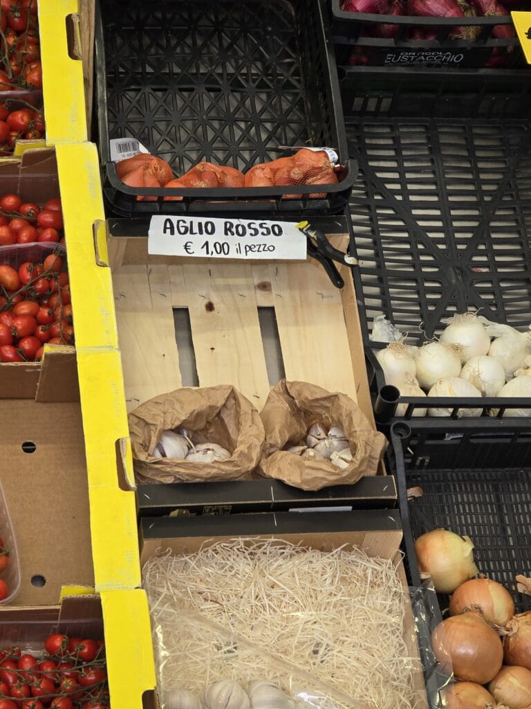 Banco di un negozio con un cartello «Aglio rosso, 1,00 € il pezzo» sopra due sacchetti di carta con teste d’aglio bianche